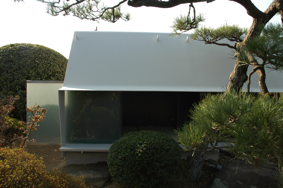 Steel Sheet Teahouse The engawa (veranda) in this minka faces the remnants of a precious Japanese-style garden, which was maintained for many years as a hobby of the preceding generation. Inheriting the shape of this bequest, a contemporary monocoque tearoom was newly installed in the garden. So as not to hurt the existing delicate garden trees, the main body of the tearoom was made in a factory, then simply placed in its location in the garden. Overall, it is divided into a half-exterior meeting part and a stoic interior space comprising an alcove and two tatami mats, covered with a roof membrane of steel sheet. An extraordinary space, this tearoom is a contemporary meditation space, and at the seasons change it is a place in dialogue with nature. As well as being also to wander through the exciting garden, visually there emerges a spatial continuity that unifies inside and outside even more than the engawa, producing a lifestyle in intimate contact with nature.<br /><br />The steel sheet that I used goes from an architectural building frame to an industrially processed product like a vehicle, and the sculptural usage avoids the impact of the strong expression of steel. The advantage of this material is its high degree of strength and the possibility of high-precision factory fabrication through using CAD; a standard quality may be maintained everywhere, the price-per-weight does not vary much, and it can cope with customization and prefabrication. The sizes for this tearoom were determined by the volume of heights and widths that are able to be transported by vehicle. It was all produced at the factory, and the 9mm-thick steel sheets were bent into curved surface at key points in order to make a unified pre-strengthened structure. Divided into two parts, it was then lowered as-it into the garden by crane. It was then reconnected, fittings were inserted, tatami mats were laid, and this was all completed over a short period of time. The inside and outside have been given a different expression. Traces of the fabrication process and welding have been left in the interior, and the outside is finished with industrial smooth painting like a vehicle, with a taste for simplicity and quiet.