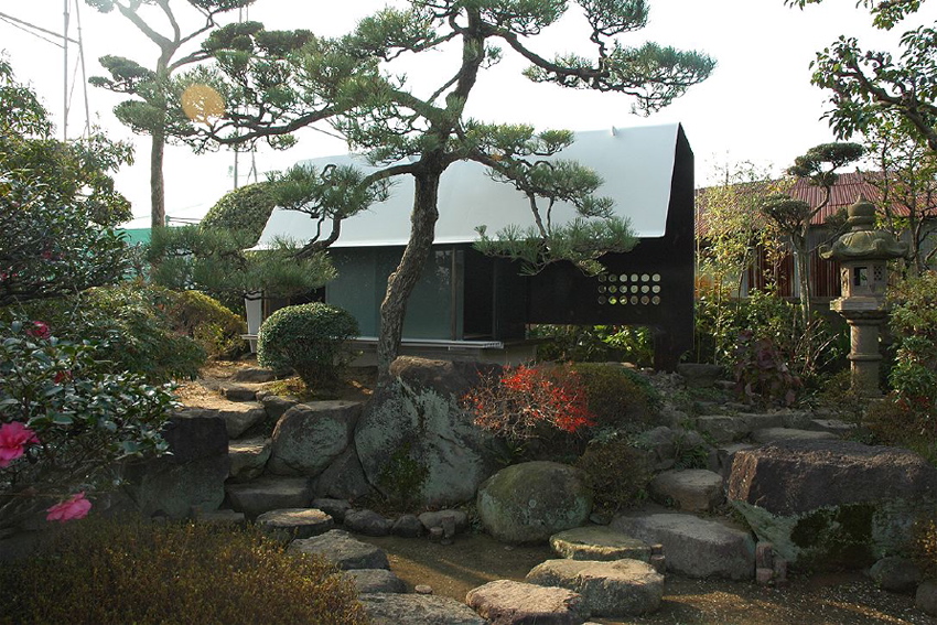 Steel Sheet Teahouse The engawa (veranda) in this minka faces the remnants of a precious Japanese-style garden, which was maintained for many years as a hobby of the preceding generation. Inheriting the shape of this bequest, a contemporary monocoque tearoom was newly installed in the garden. So as not to hurt the existing delicate garden trees, the main body of the tearoom was made in a factory, then simply placed in its location in the garden. Overall, it is divided into a half-exterior meeting part and a stoic interior space comprising an alcove and two tatami mats, covered with a roof membrane of steel sheet. An extraordinary space, this tearoom is a contemporary meditation space, and at the seasons change it is a place in dialogue with nature. As well as being also to wander through the exciting garden, visually there emerges a spatial continuity that unifies inside and outside even more than the engawa, producing a lifestyle in intimate contact with nature.<br /><br />The steel sheet that I used goes from an architectural building frame to an industrially processed product like a vehicle, and the sculptural usage avoids the impact of the strong expression of steel. The advantage of this material is its high degree of strength and the possibility of high-precision factory fabrication through using CAD; a standard quality may be maintained everywhere, the price-per-weight does not vary much, and it can cope with customization and prefabrication. The sizes for this tearoom were determined by the volume of heights and widths that are able to be transported by vehicle. It was all produced at the factory, and the 9mm-thick steel sheets were bent into curved surface at key points in order to make a unified pre-strengthened structure. Divided into two parts, it was then lowered as-it into the garden by crane. It was then reconnected, fittings were inserted, tatami mats were laid, and this was all completed over a short period of time. The inside and outside have been given a different expression. Traces of the fabrication process and welding have been left in the interior, and the outside is finished with industrial smooth painting like a vehicle, with a taste for simplicity and quiet.