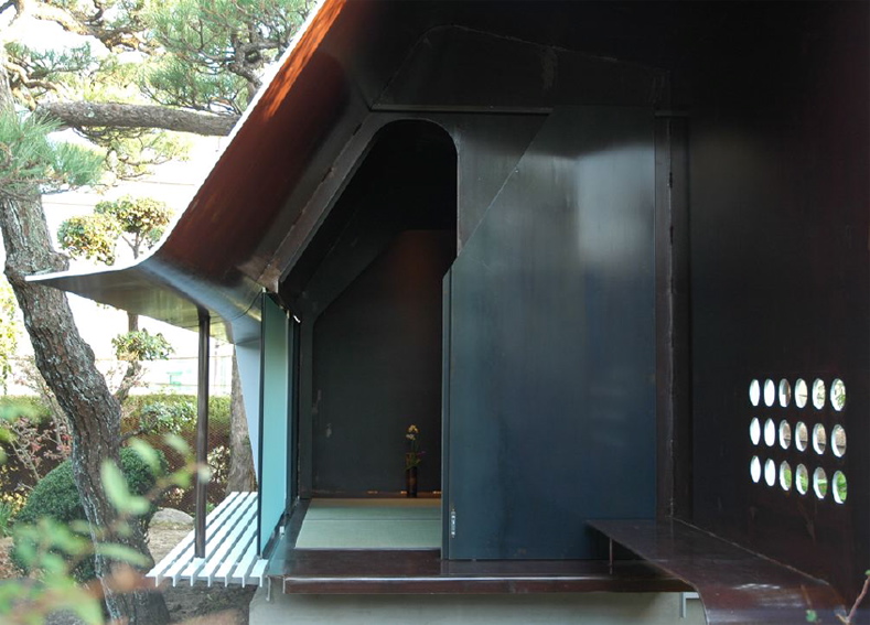 Steel Sheet Teahouse The engawa (veranda) in this minka faces the remnants of a precious Japanese-style garden, which was maintained for many years as a hobby of the preceding generation. Inheriting the shape of this bequest, a contemporary monocoque tearoom was newly installed in the garden. So as not to hurt the existing delicate garden trees, the main body of the tearoom was made in a factory, then simply placed in its location in the garden. Overall, it is divided into a half-exterior meeting part and a stoic interior space comprising an alcove and two tatami mats, covered with a roof membrane of steel sheet. An extraordinary space, this tearoom is a contemporary meditation space, and at the seasons change it is a place in dialogue with nature. As well as being also to wander through the exciting garden, visually there emerges a spatial continuity that unifies inside and outside even more than the engawa, producing a lifestyle in intimate contact with nature.<br /><br />The steel sheet that I used goes from an architectural building frame to an industrially processed product like a vehicle, and the sculptural usage avoids the impact of the strong expression of steel. The advantage of this material is its high degree of strength and the possibility of high-precision factory fabrication through using CAD; a standard quality may be maintained everywhere, the price-per-weight does not vary much, and it can cope with customization and prefabrication. The sizes for this tearoom were determined by the volume of heights and widths that are able to be transported by vehicle. It was all produced at the factory, and the 9mm-thick steel sheets were bent into curved surface at key points in order to make a unified pre-strengthened structure. Divided into two parts, it was then lowered as-it into the garden by crane. It was then reconnected, fittings were inserted, tatami mats were laid, and this was all completed over a short period of time. The inside and outside have been given a different expression. Traces of the fabrication process and welding have been left in the interior, and the outside is finished with industrial smooth painting like a vehicle, with a taste for simplicity and quiet.