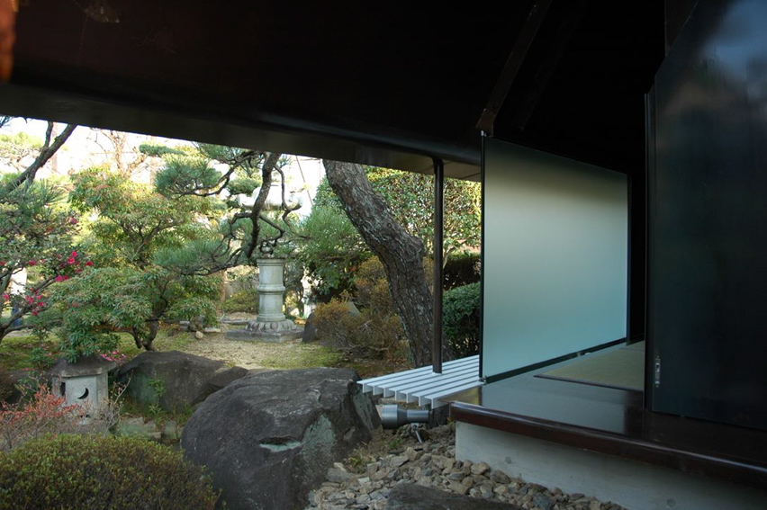 Steel Sheet Teahouse The engawa (veranda) in this minka faces the remnants of a precious Japanese-style garden, which was maintained for many years as a hobby of the preceding generation. Inheriting the shape of this bequest, a contemporary monocoque tearoom was newly installed in the garden. So as not to hurt the existing delicate garden trees, the main body of the tearoom was made in a factory, then simply placed in its location in the garden. Overall, it is divided into a half-exterior meeting part and a stoic interior space comprising an alcove and two tatami mats, covered with a roof membrane of steel sheet. An extraordinary space, this tearoom is a contemporary meditation space, and at the seasons change it is a place in dialogue with nature. As well as being also to wander through the exciting garden, visually there emerges a spatial continuity that unifies inside and outside even more than the engawa, producing a lifestyle in intimate contact with nature.<br /><br />The steel sheet that I used goes from an architectural building frame to an industrially processed product like a vehicle, and the sculptural usage avoids the impact of the strong expression of steel. The advantage of this material is its high degree of strength and the possibility of high-precision factory fabrication through using CAD; a standard quality may be maintained everywhere, the price-per-weight does not vary much, and it can cope with customization and prefabrication. The sizes for this tearoom were determined by the volume of heights and widths that are able to be transported by vehicle. It was all produced at the factory, and the 9mm-thick steel sheets were bent into curved surface at key points in order to make a unified pre-strengthened structure. Divided into two parts, it was then lowered as-it into the garden by crane. It was then reconnected, fittings were inserted, tatami mats were laid, and this was all completed over a short period of time. The inside and outside have been given a different expression. Traces of the fabrication process and welding have been left in the interior, and the outside is finished with industrial smooth painting like a vehicle, with a taste for simplicity and quiet.